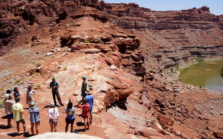 A group of people stand in a circle on red rocks high above a winding river.